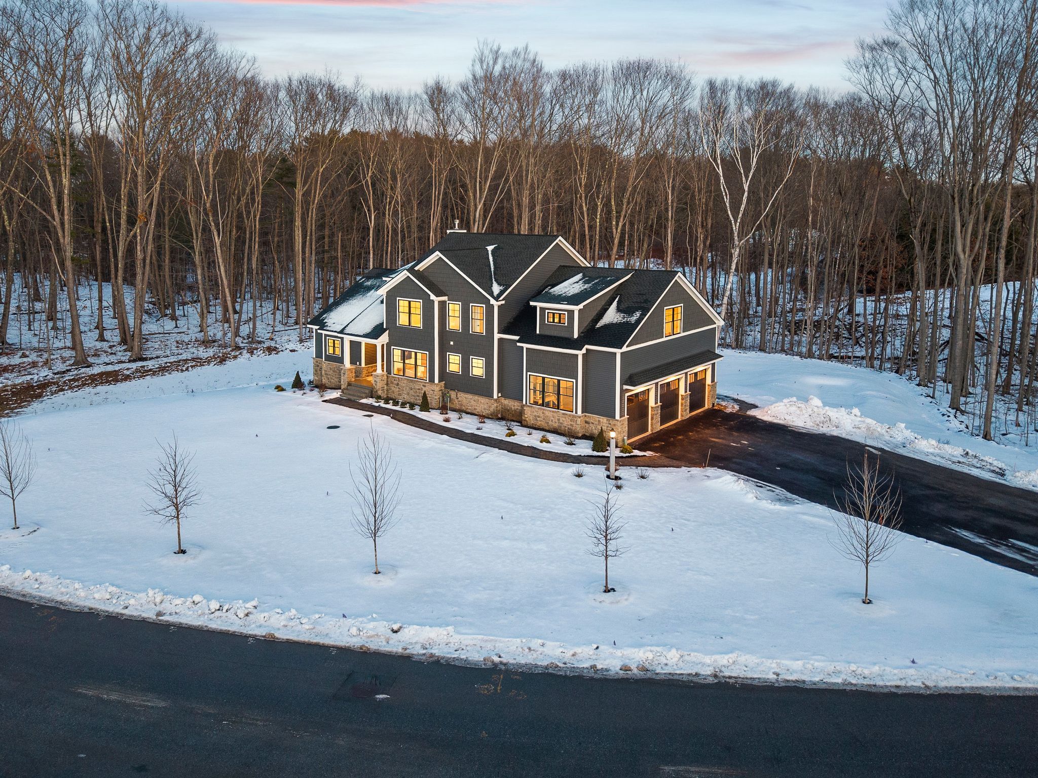 Modern black house with large windows, snow-covered front yard, surrounded by trees, in winter season, aerial view.