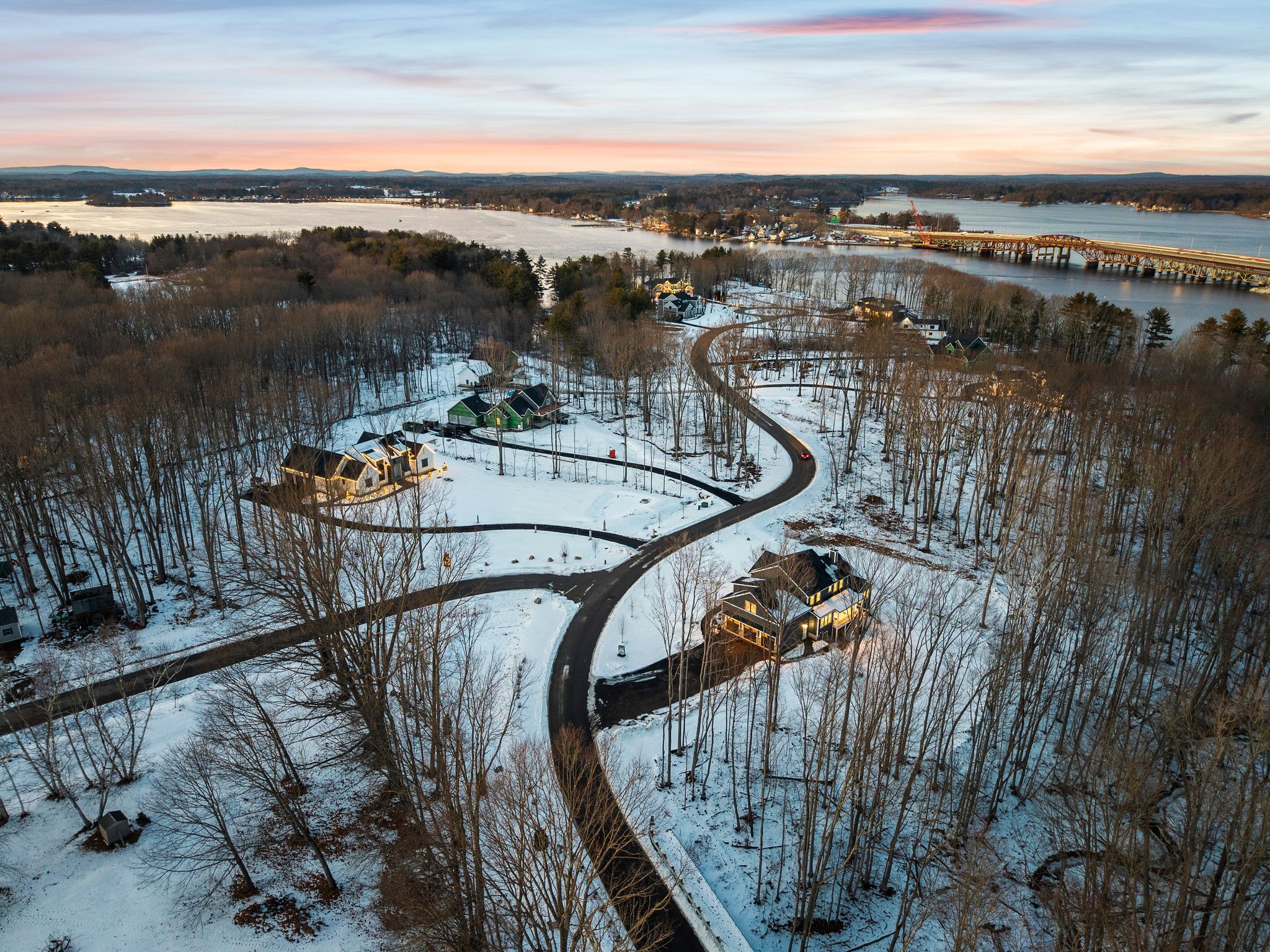 Aerial view of snow-covered houses along a winding road, surrounded by leafless trees and overlooking a river with a bridge at sunset.