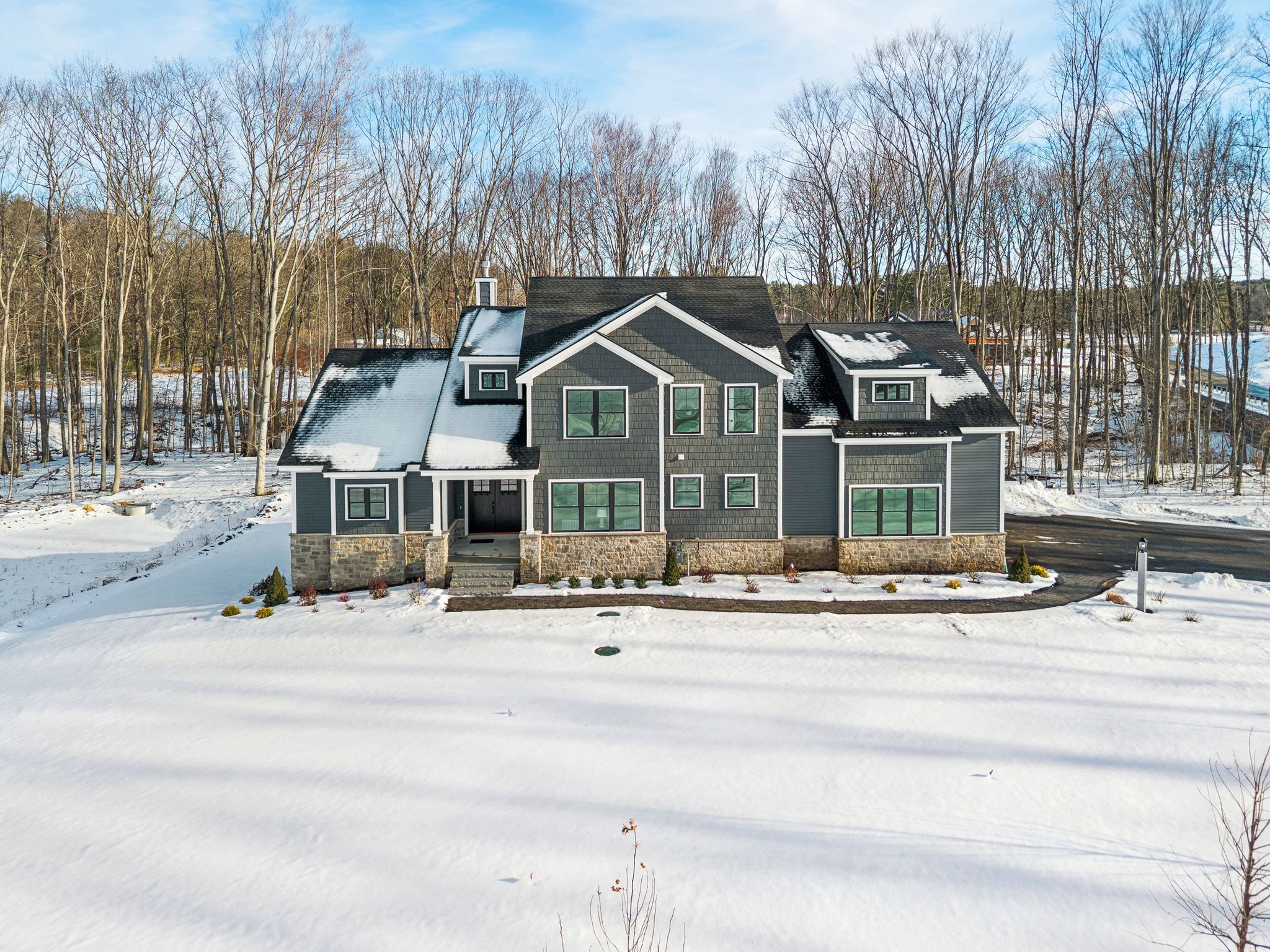 Modern gray and black two-story house with stone accents on foundation surrounded by snow and trees in winter.
