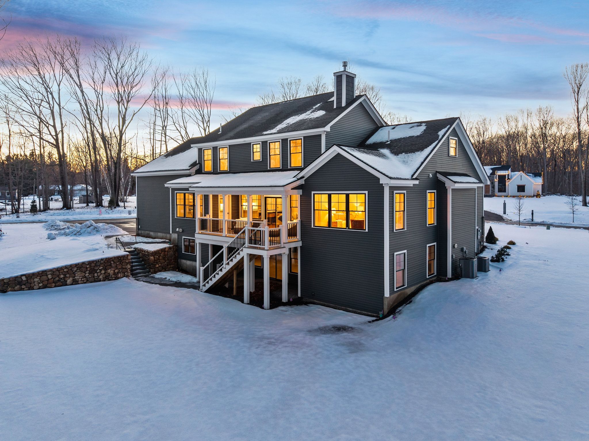 Spacious dark gray two-story house with warm yellow interior lighting, surrounded by snow-covered yard during winter sunset, with leafless trees in the background.