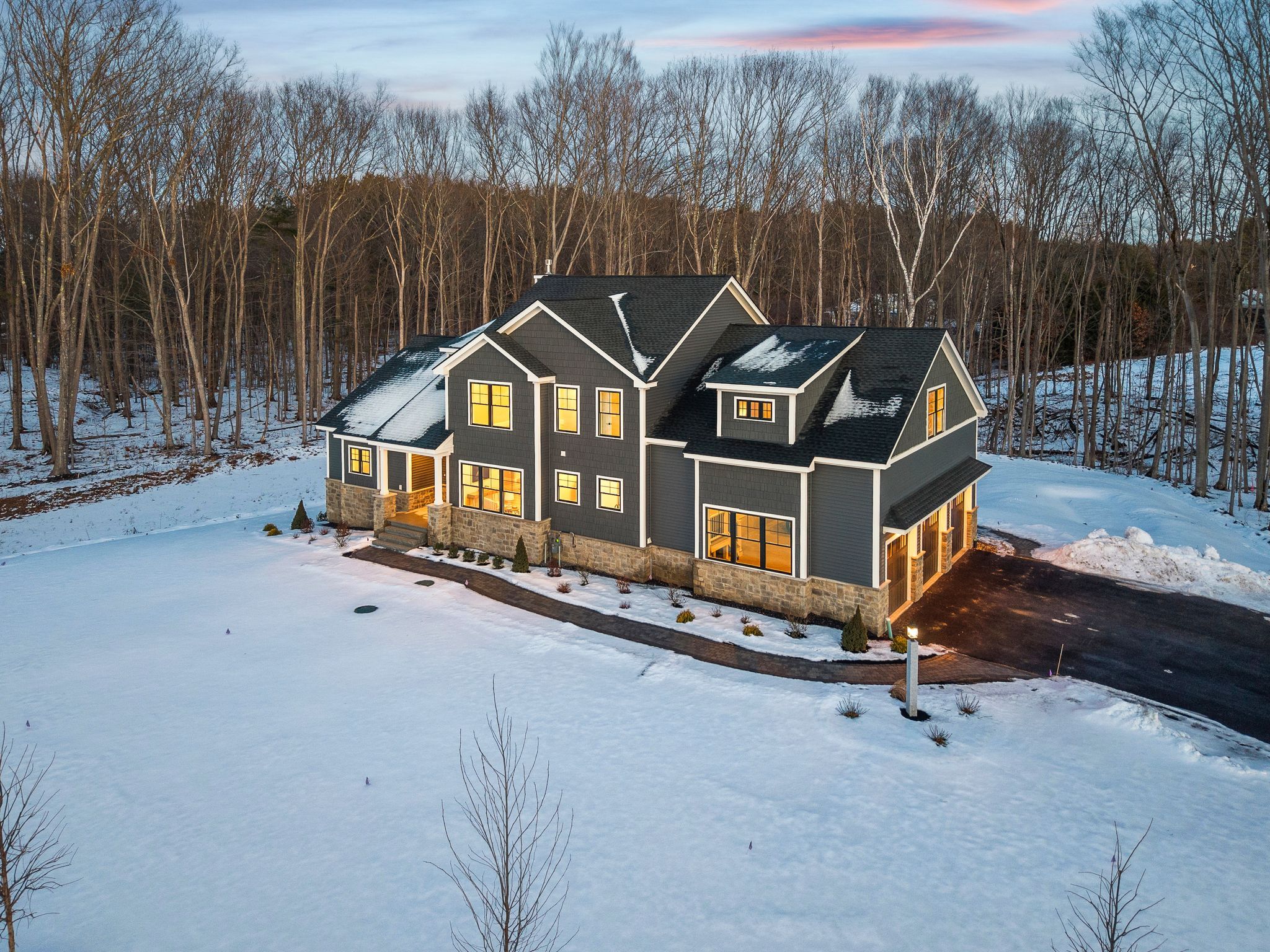 Modern contemporary house with large windows, stone and gray siding, surrounded by snow, located near a wooded area during sunset.