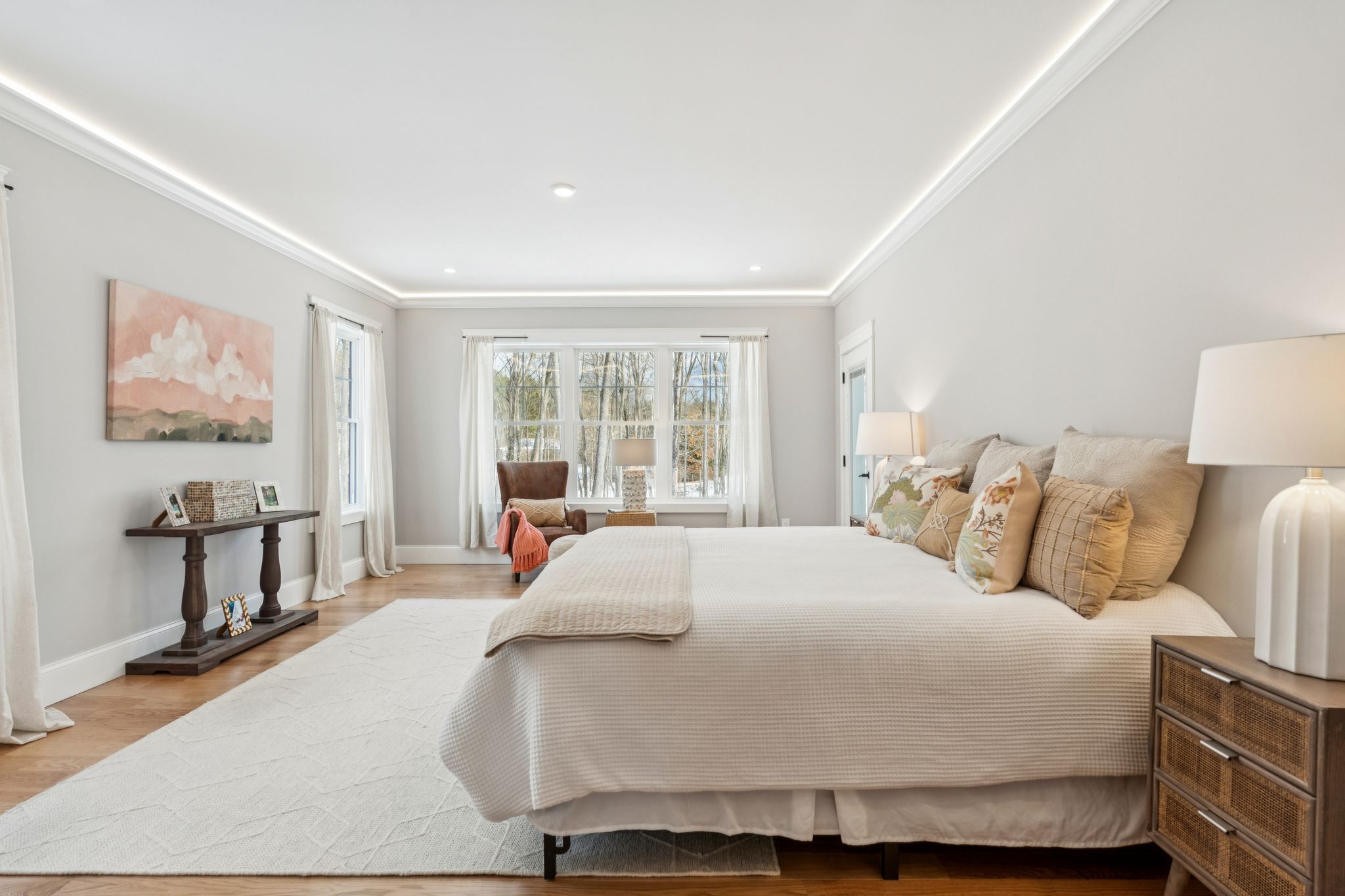 Bright and airy bedroom with large window, white walls, beige bedding, wooden furniture, and decorative pillows.