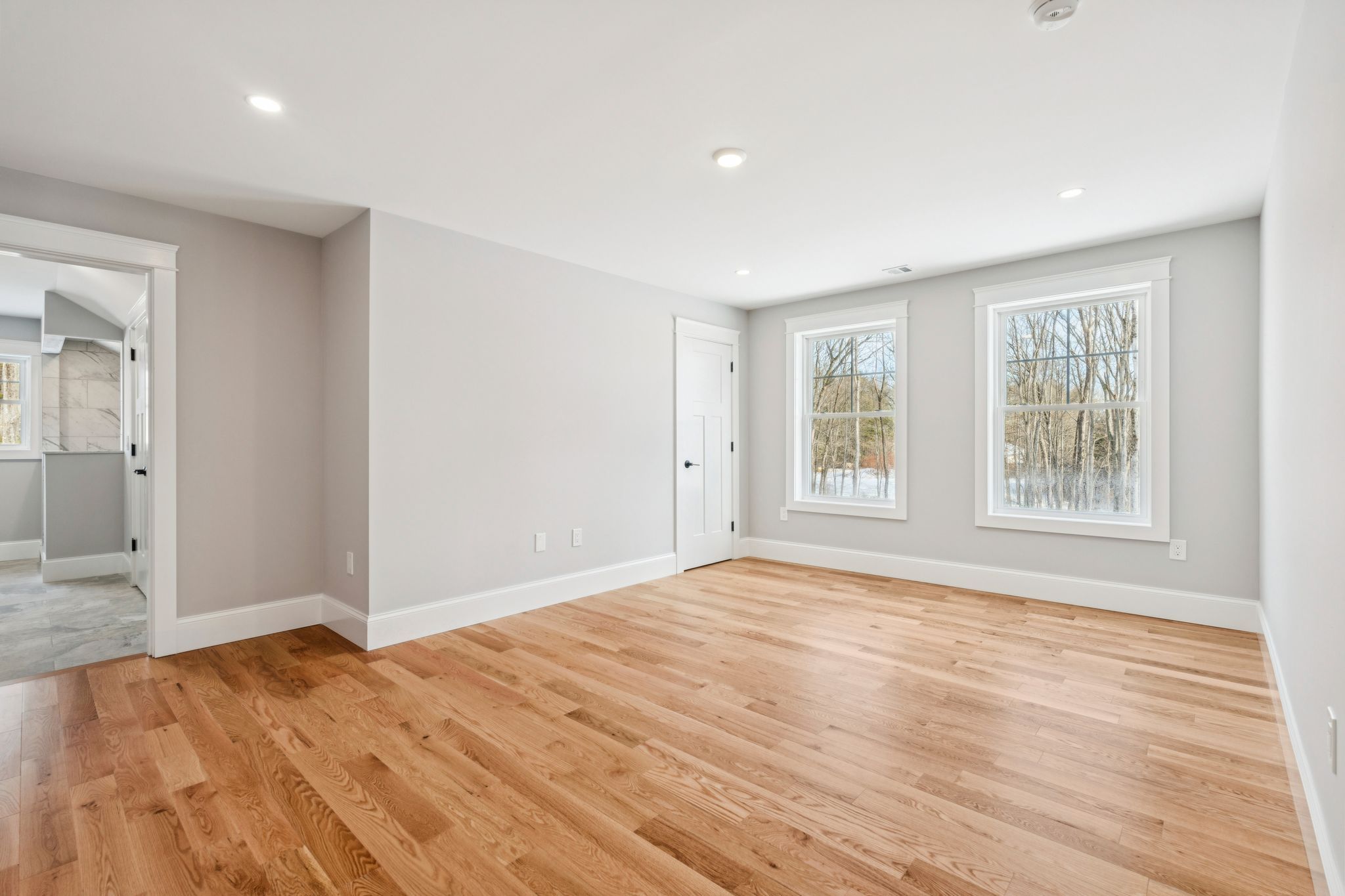 Bright empty living room with natural light, hardwood flooring, white walls, and large windows showing trees outside.