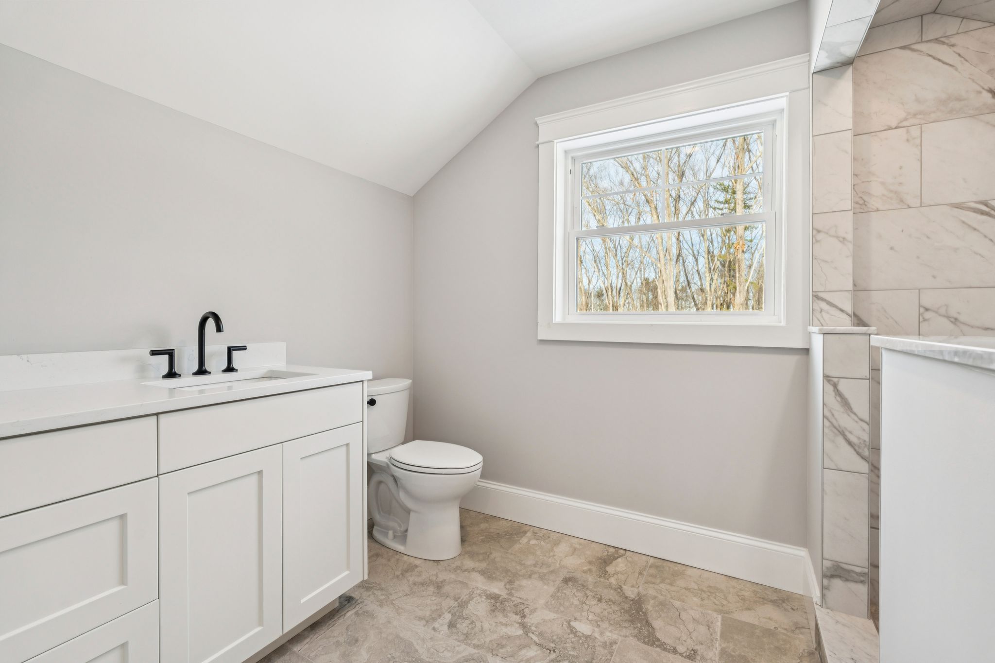 Modern minimalist bathroom with white vanity, black fixtures, toilet, large window, and marble accents