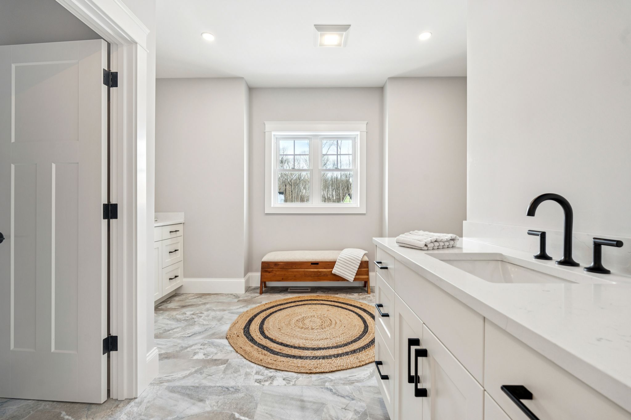 Bright and modern bathroom with white cabinetry, black fixtures, a round woven rug, and a window overlooking trees.