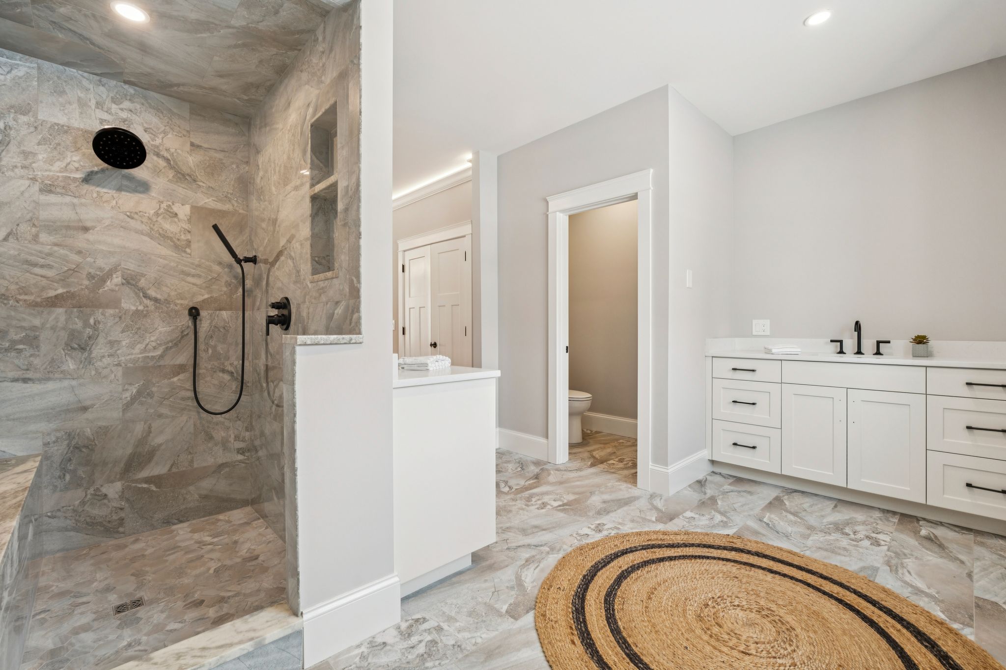 Modern bathroom with walk-in shower featuring gray marble tiles, white vanity with black hardware, and a separate toilet area with doorway, beige and gray tiled flooring, and a round jute rug.