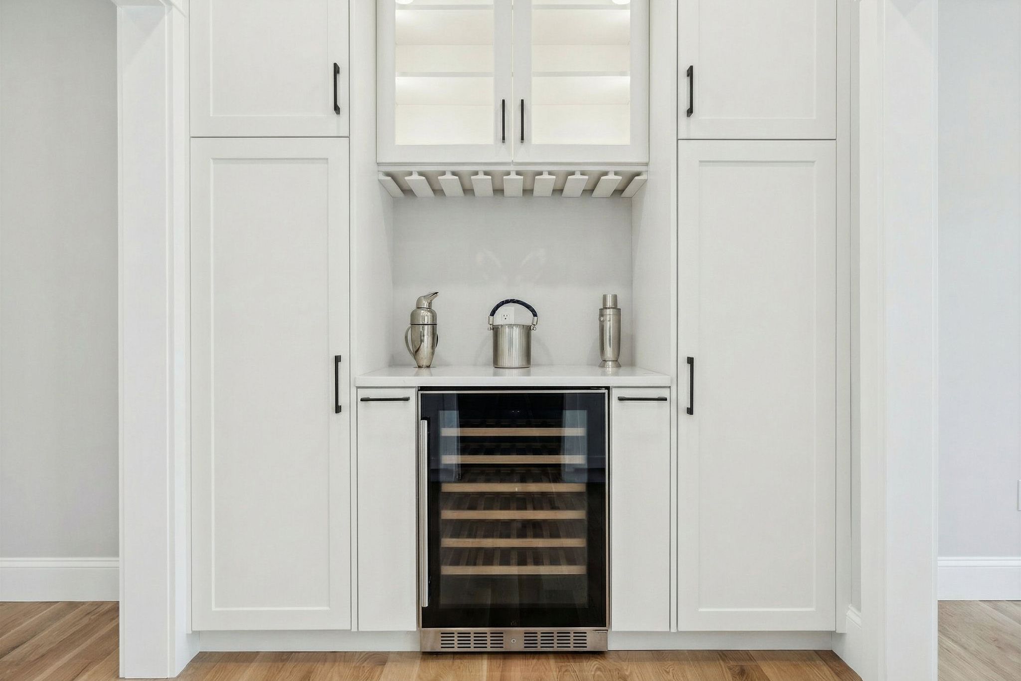 Modern wine storage area with a built-in wine cooler, white cabinetry, stainless steel wine accessories, and a minimalist design.