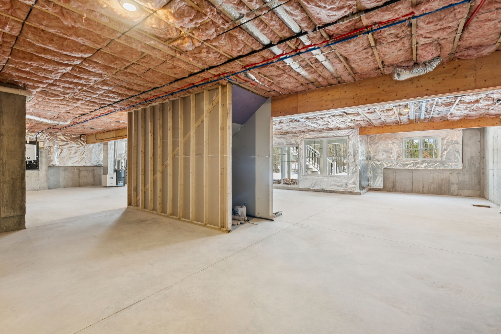 Interior view of a house under construction with exposed insulation, unfinished walls, and large windows, showing an open-concept layout with concrete flooring and partial framing.