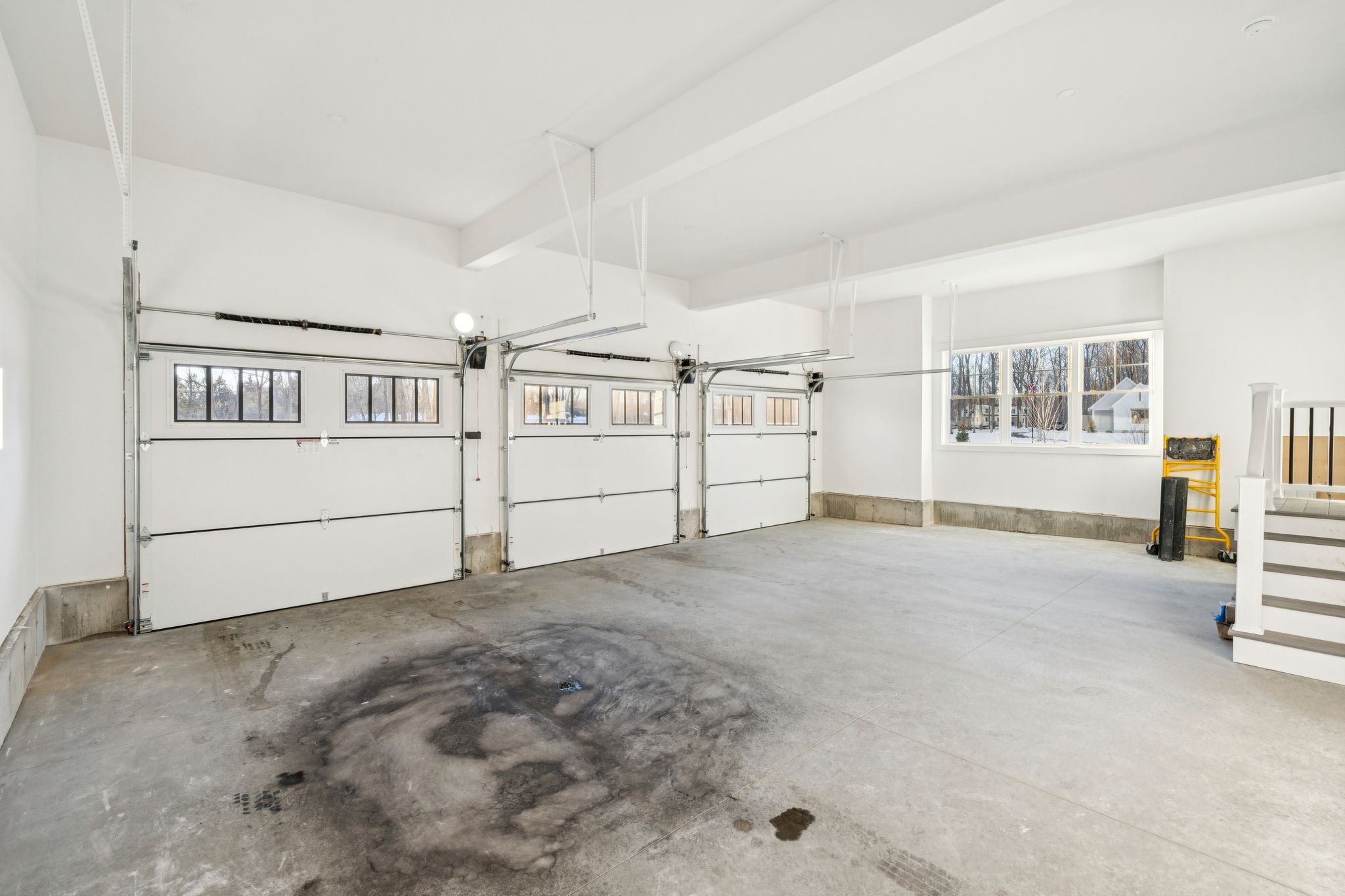 Empty modern two-car garage with white walls, concrete floor with oil stains, large window, and storage area with stairs
