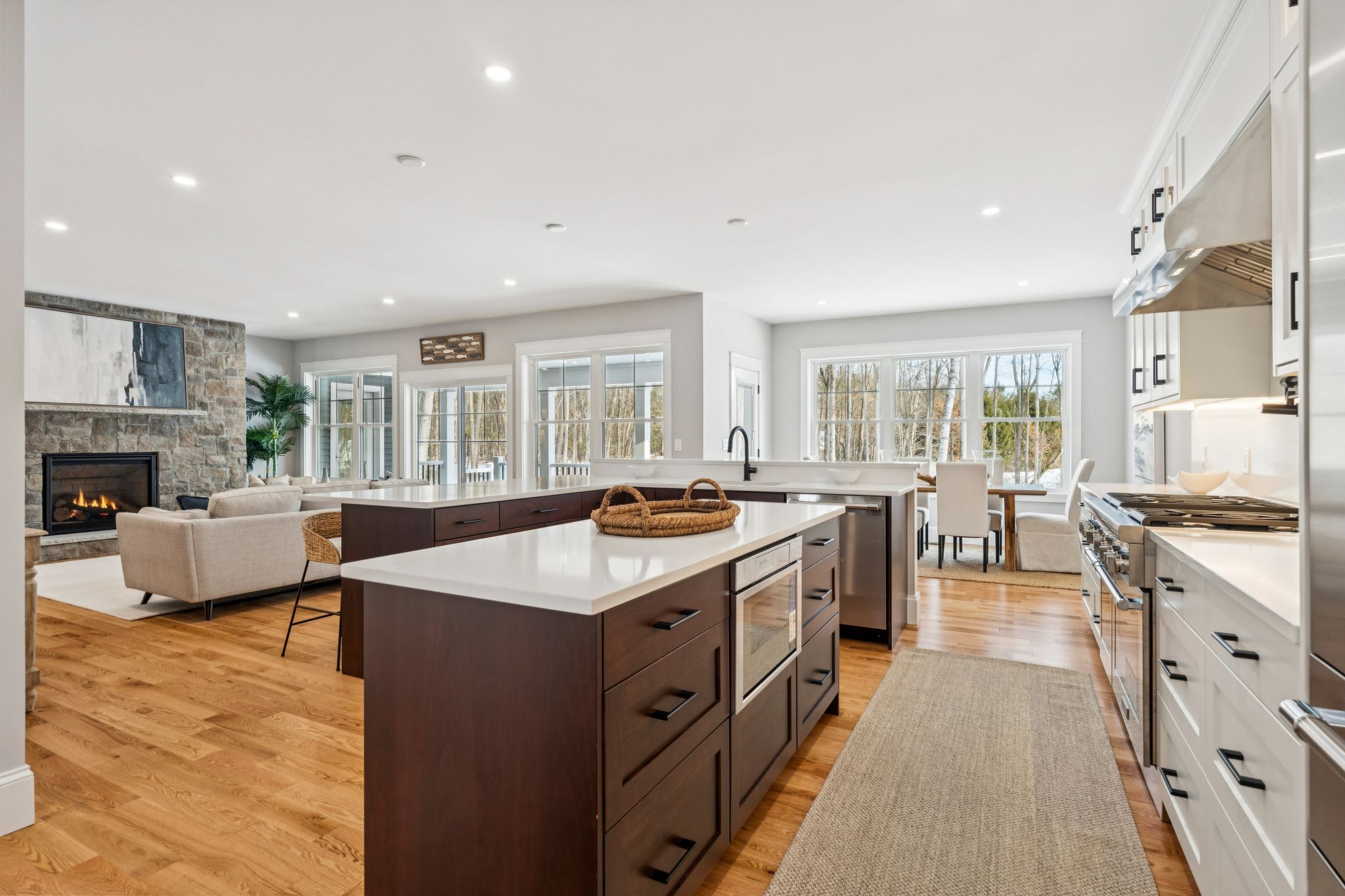 Bright, open-concept living room and kitchen with large windows overlooking a wooded outdoor view, featuring a modern kitchen island with dark wood cabinetry and white countertops, a cozy living area with a stone fireplace, and stylish dining space.