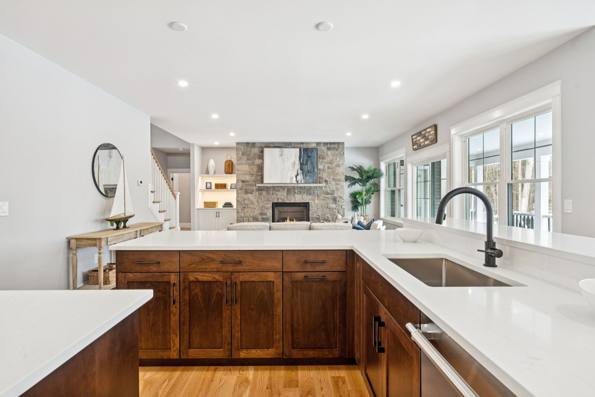Open-concept kitchen with white countertops, dark wooden cabinets, stainless steel sink and faucet, overlooking a spacious living room with stone fireplace, framed artwork, large windows, and a cozy seating area with a potted plant.