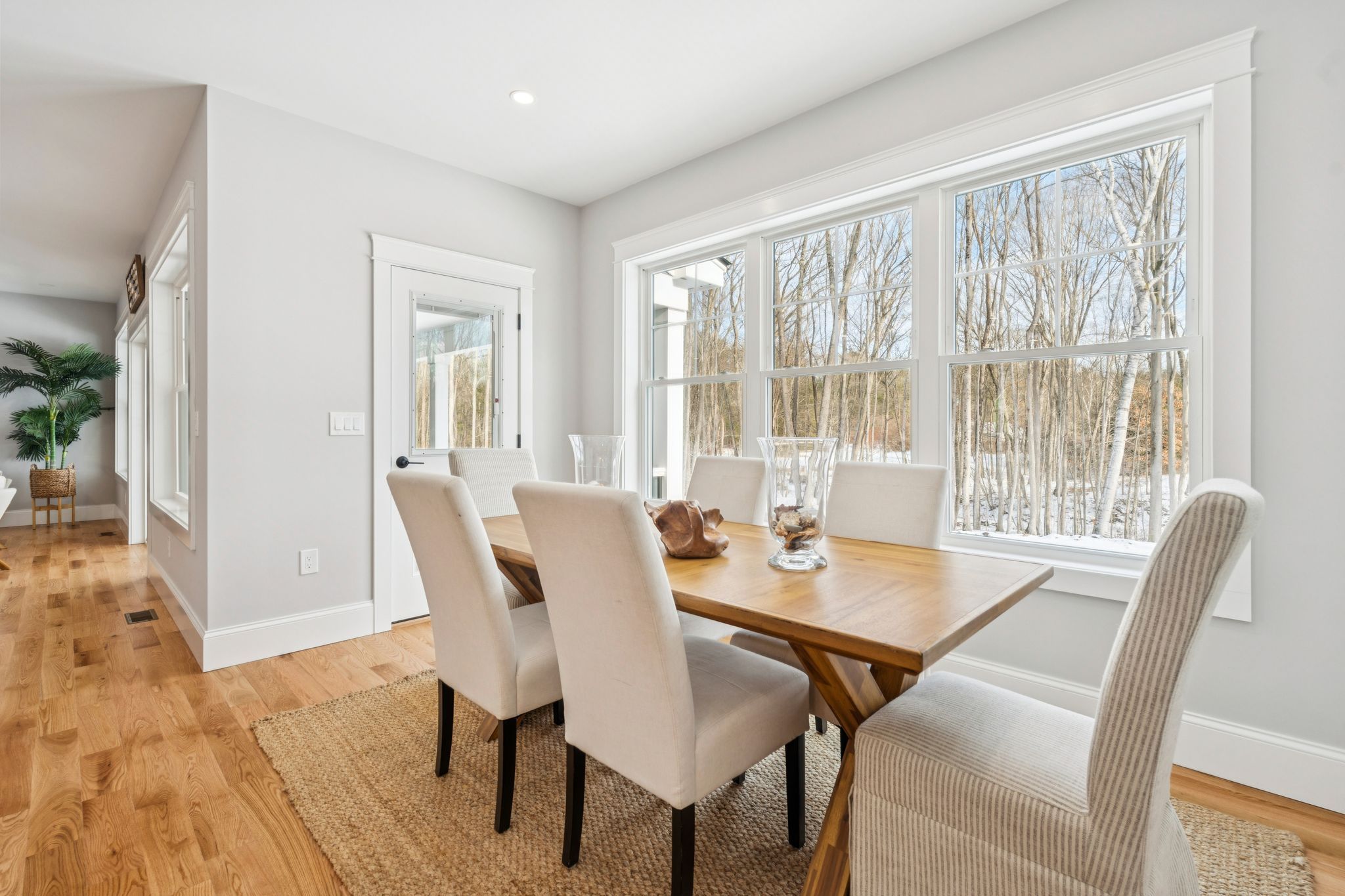 Bright dining room with large windows overlooking a wooded outdoor area, featuring a wooden table and six upholstered chairs, natural light, and hardwood flooring.