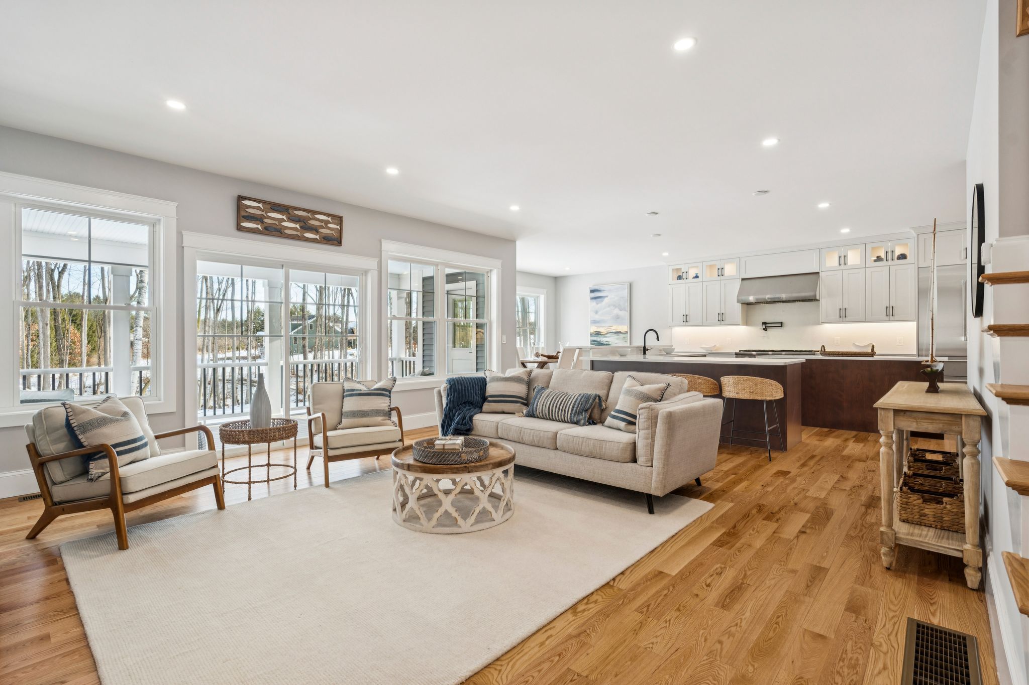 Bright open-concept living room with large windows overlooking a snowy landscape, featuring a beige sofa, wooden armchairs with cushions, a rustic coffee table, and a view into a modern kitchen with white cabinetry and a central island