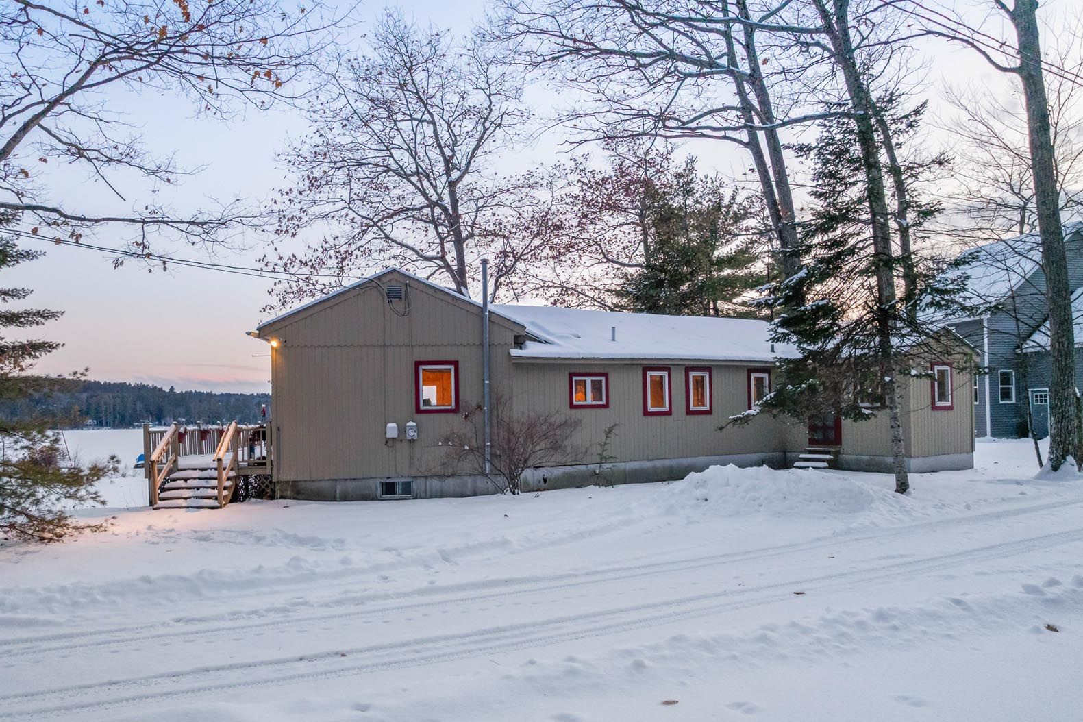 A lakeside house surrounded by lush green trees with a small dock and two colorful Adirondack chairs on the grassy shore.