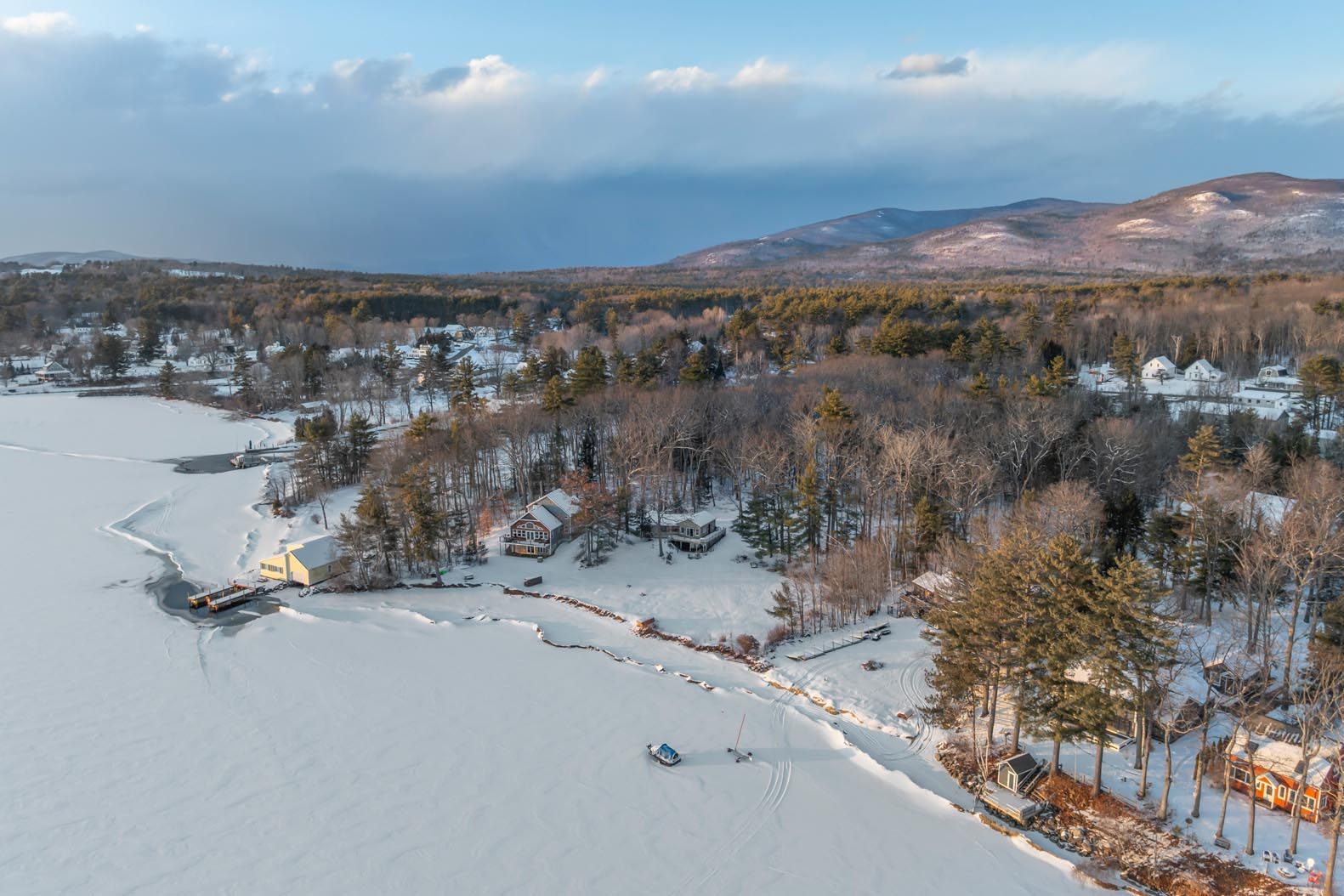 Scenic lakeside view with green grass, colorful Adirondack chairs, a dock, boats on calm water, lush tree-lined shoreline, blue sky, and distant mountains.