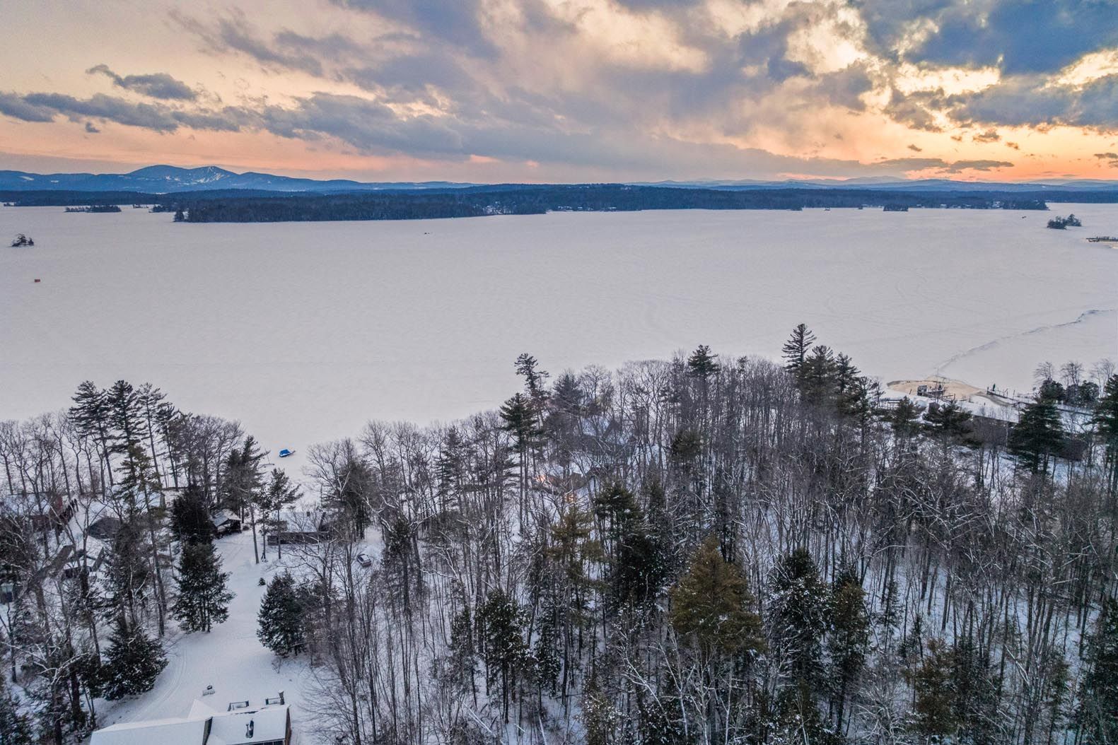 Scenic lakeside view with grassy shore, colorful lawn chairs, boats on calm water, and lush green trees under a clear blue sky.