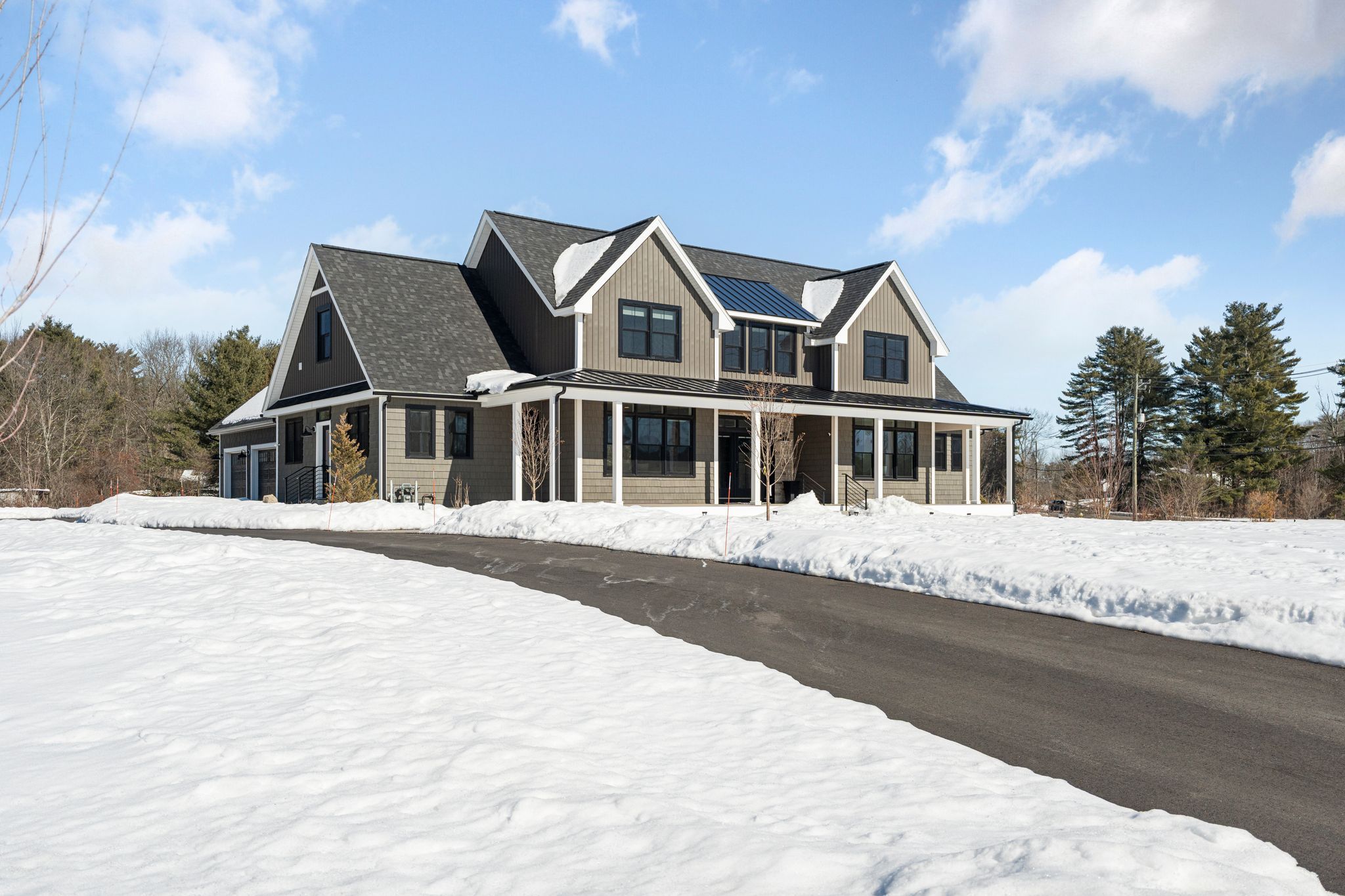 Aerial view of a snowy residential area near a river with a bridge in the background and a winding driveway leading to houses surrounded by leafless trees.