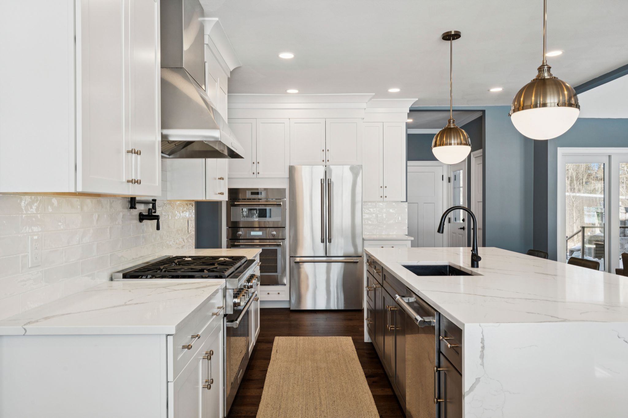 Aerial view of a snowy residential area near a river with a bridge in the background and a winding driveway leading to houses surrounded by leafless trees.