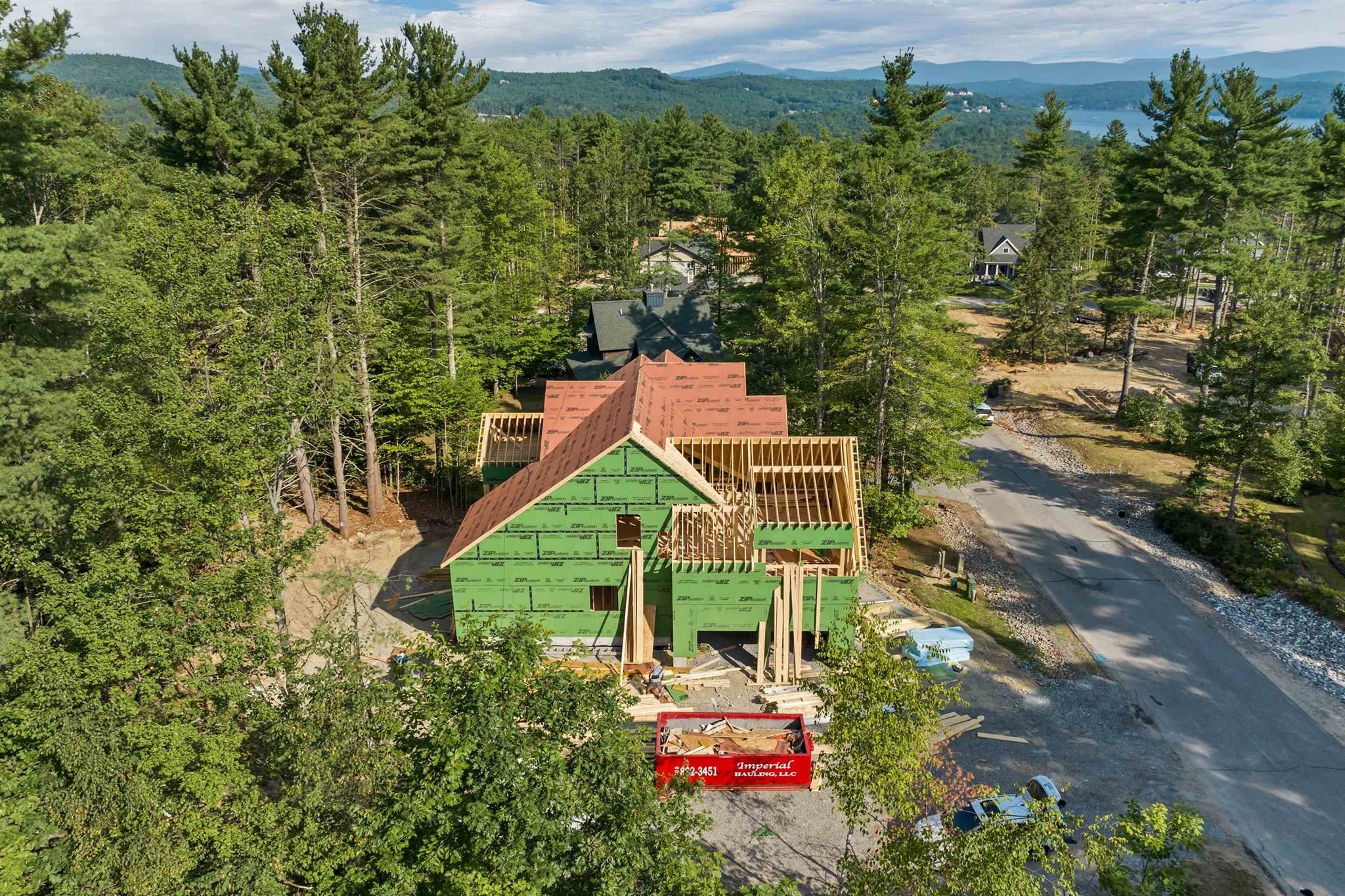 A house under construction in a wooded area with trees, a gravel driveway, and mountains in the background.
