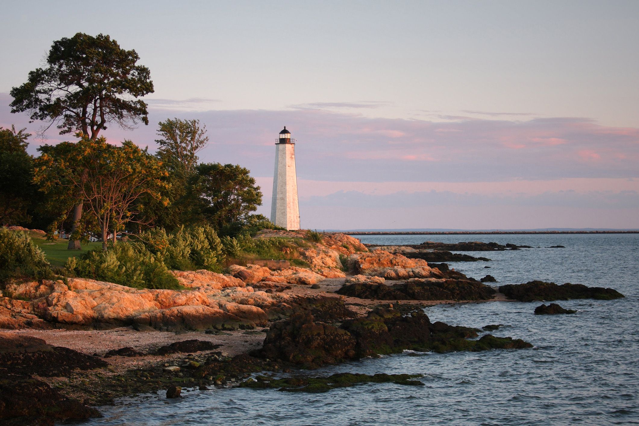 Lighthouse on a rocky coastline at sunset with trees and calm ocean water.