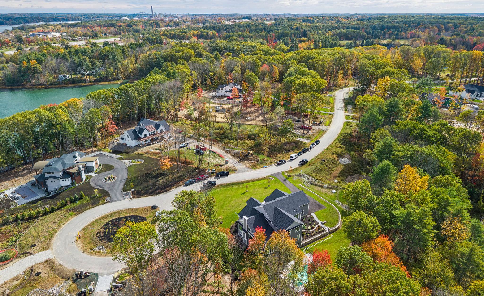 Aerial view of a suburban neighborhood surrounded by colorful autumn trees, featuring a winding road, modern houses, a lake, and distant city skyline.