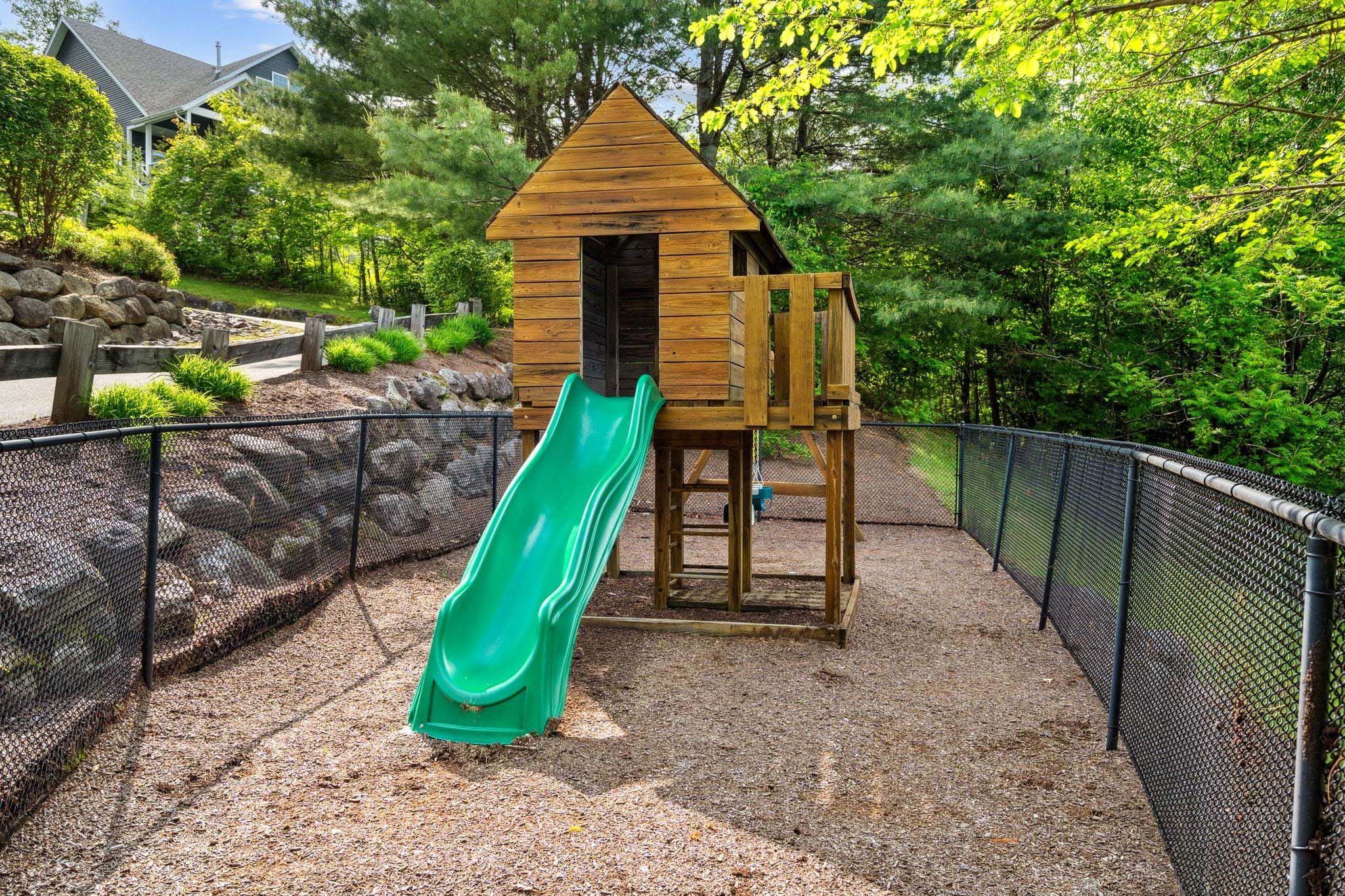 Cozy home office with a daybed, plaid bedding, front and window-side desks with monitors, white cabinets, and a patterned rug in a room with green walls and large windows overlooking a lush outdoor area.