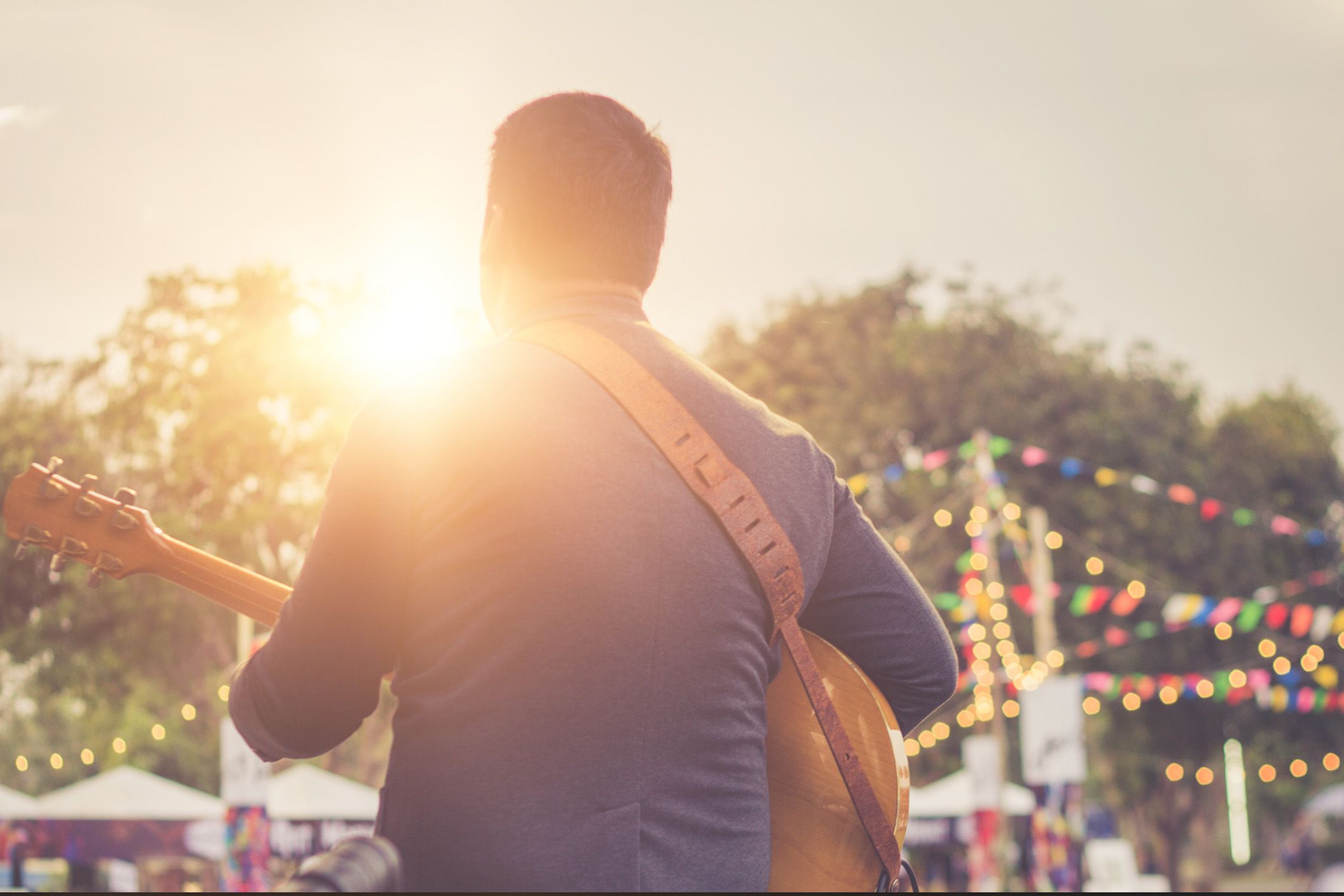 Man playing acoustic guitar outdoors at sunset during a festival with colorful string lights and trees in the background