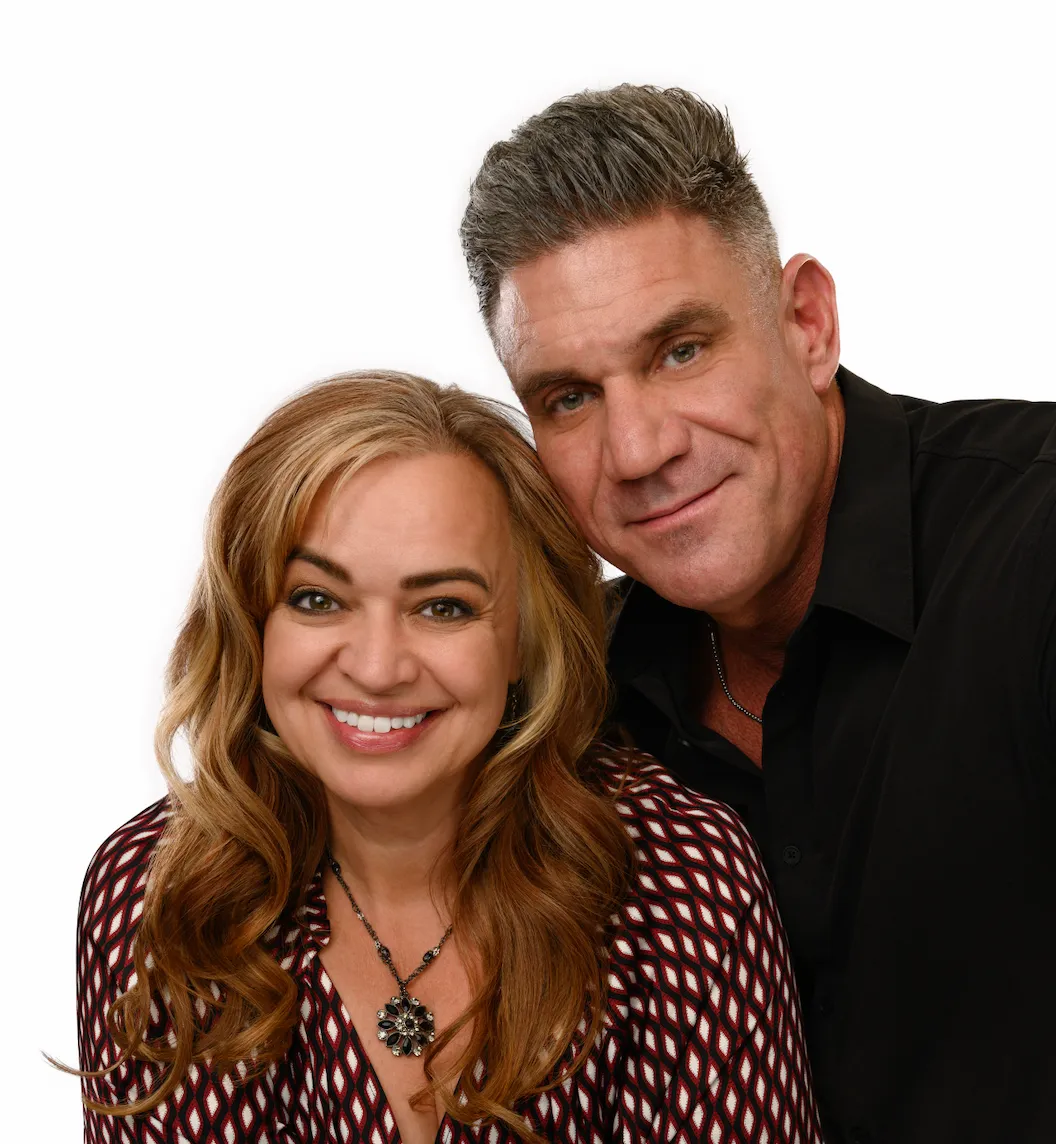 Smiling woman with long red hair wearing a patterned blouse and a necklace, standing next to a smiling man with short gray hair wearing a black shirt, against a white background