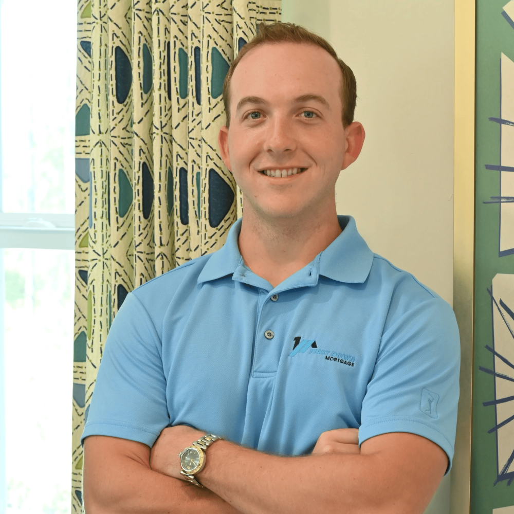 Professional portrait of a smiling bald man with a beard wearing a dark blue suit and light blue shirt, standing in front of a 
