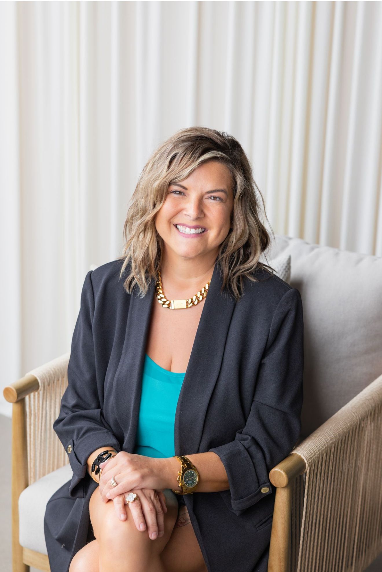 Portrait of a smiling woman with medium, wavy blonde hair wearing a black blazer, turquoise top, and gold jewelry against a light gray background.