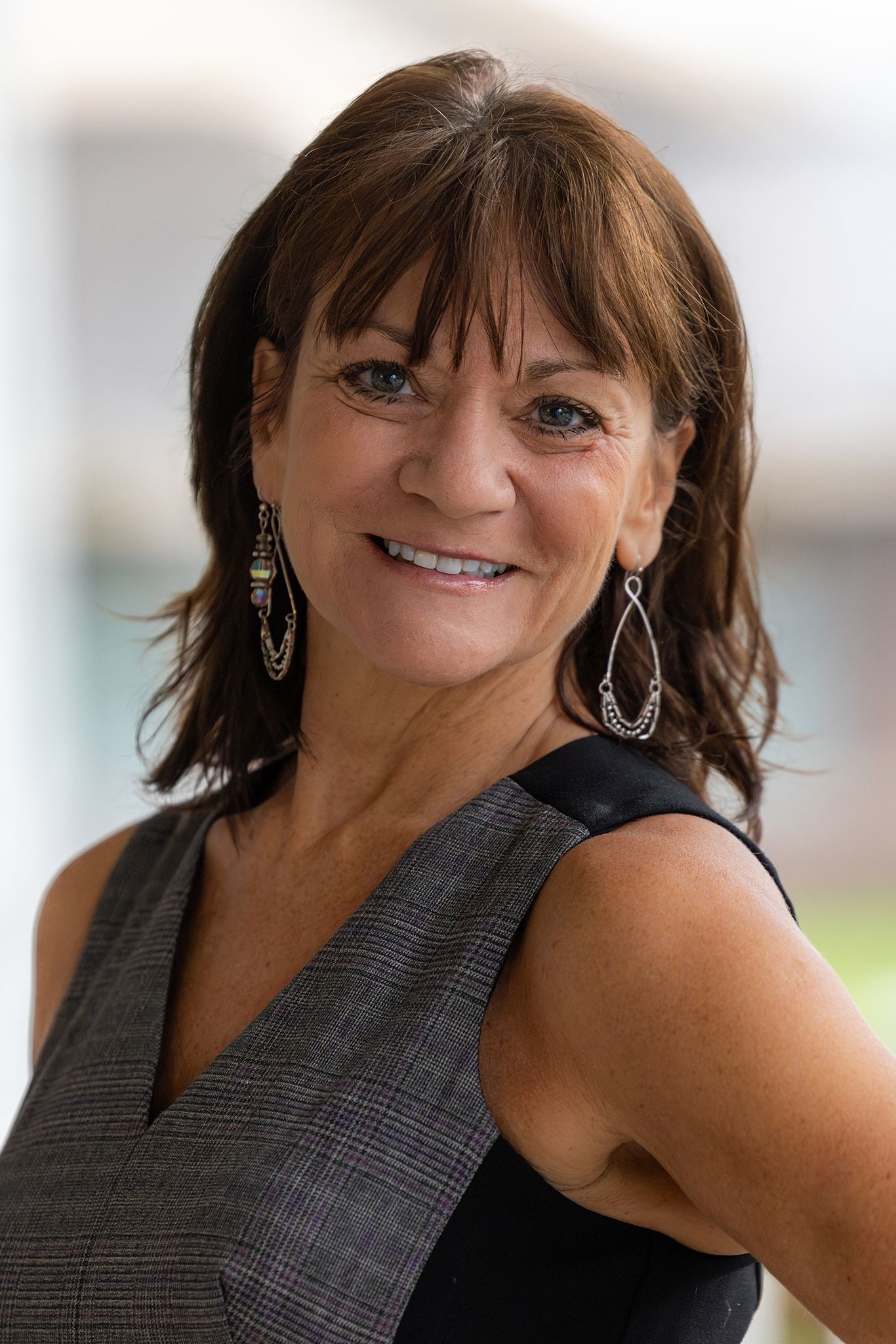 A smiling woman with medium, brown wavy hair, wearing a black and gray, sleeveless top, one gold looped earring, and one silver looped earring.