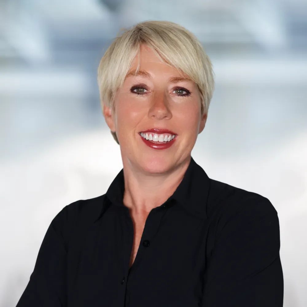 Happy professional woman with blonde hair smiling, wearing a black blazer and white shirt, in a modern office environment.