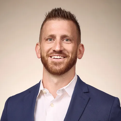 Smiling man in a navy suit and white patterned shirt, professional headshot against plain background