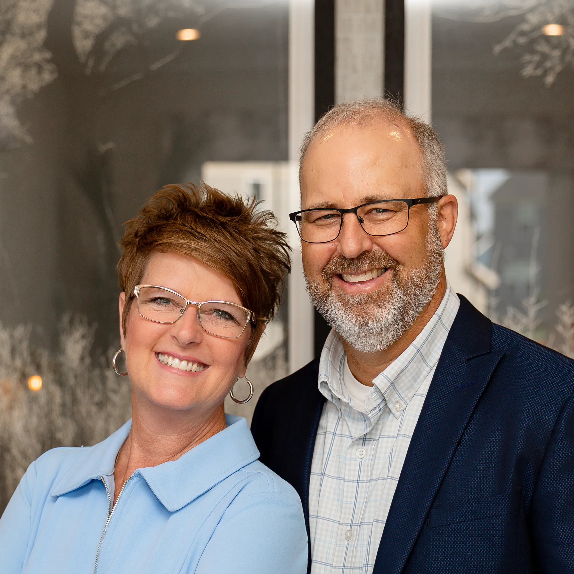 Smiling couple in professional attire standing together against a plain background, ?? and ??, wearing glasses, woman with short hair and earrings, man with a beard and receding hairline, in black shirts, black and white photo