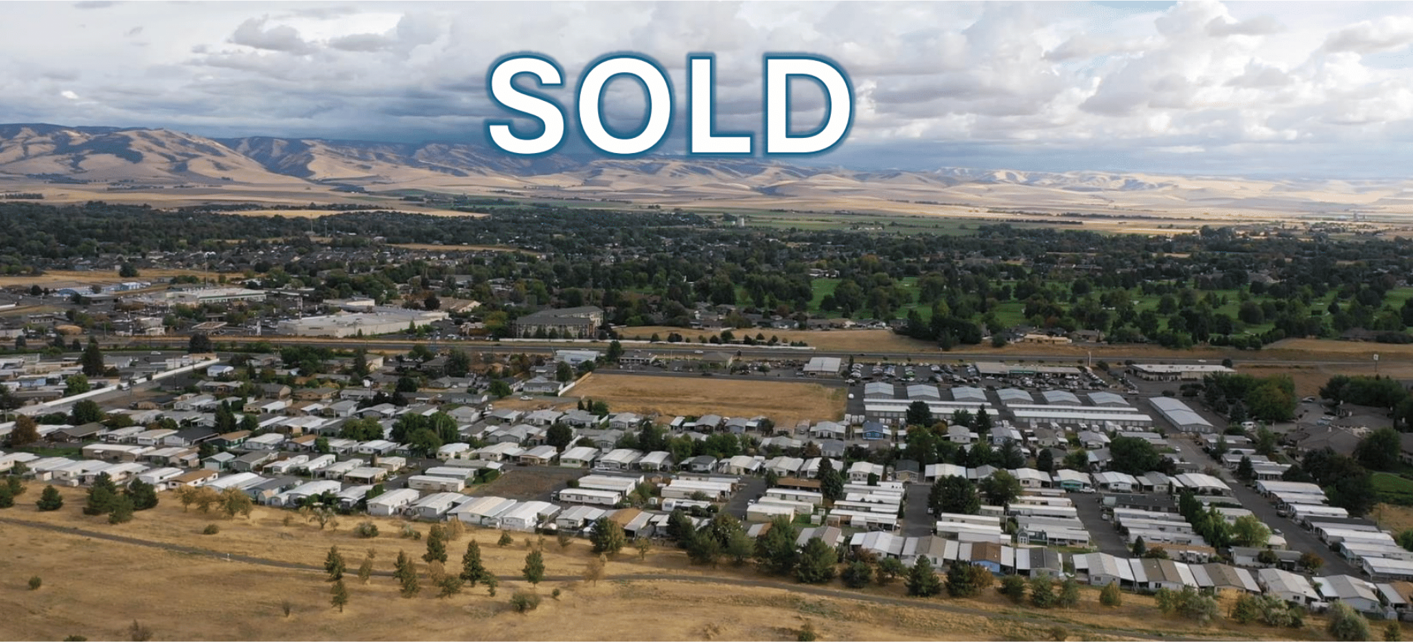 Aerial view of a suburban neighborhood with mobile homes and small houses, surrounded by green and dry fields, with mountains in the background, overlaid with the large blue and white text 