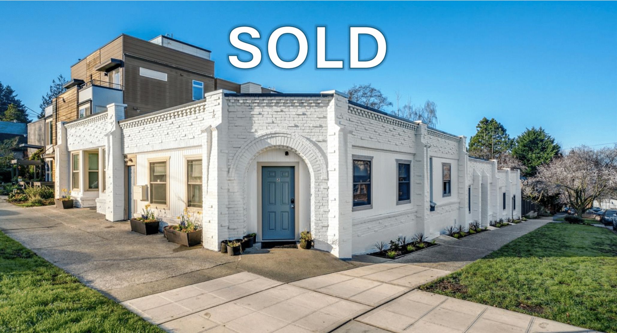 Sold sign on a white brick residential building in a suburban neighborhood with manicured lawn and flowering trees.