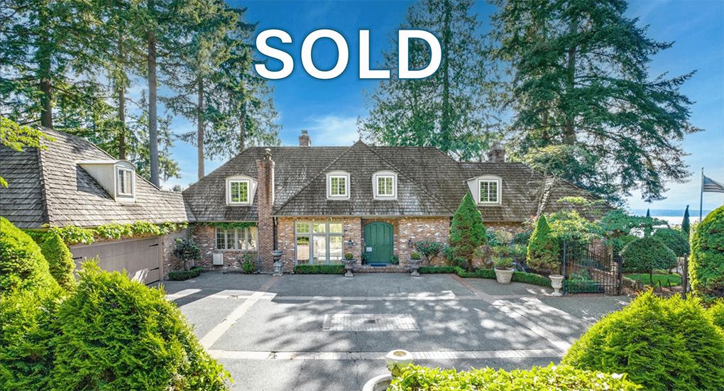 Sold sign on a house with a brick exterior, green door, and lush landscaping in a suburban setting with trees and a view of water in the background.