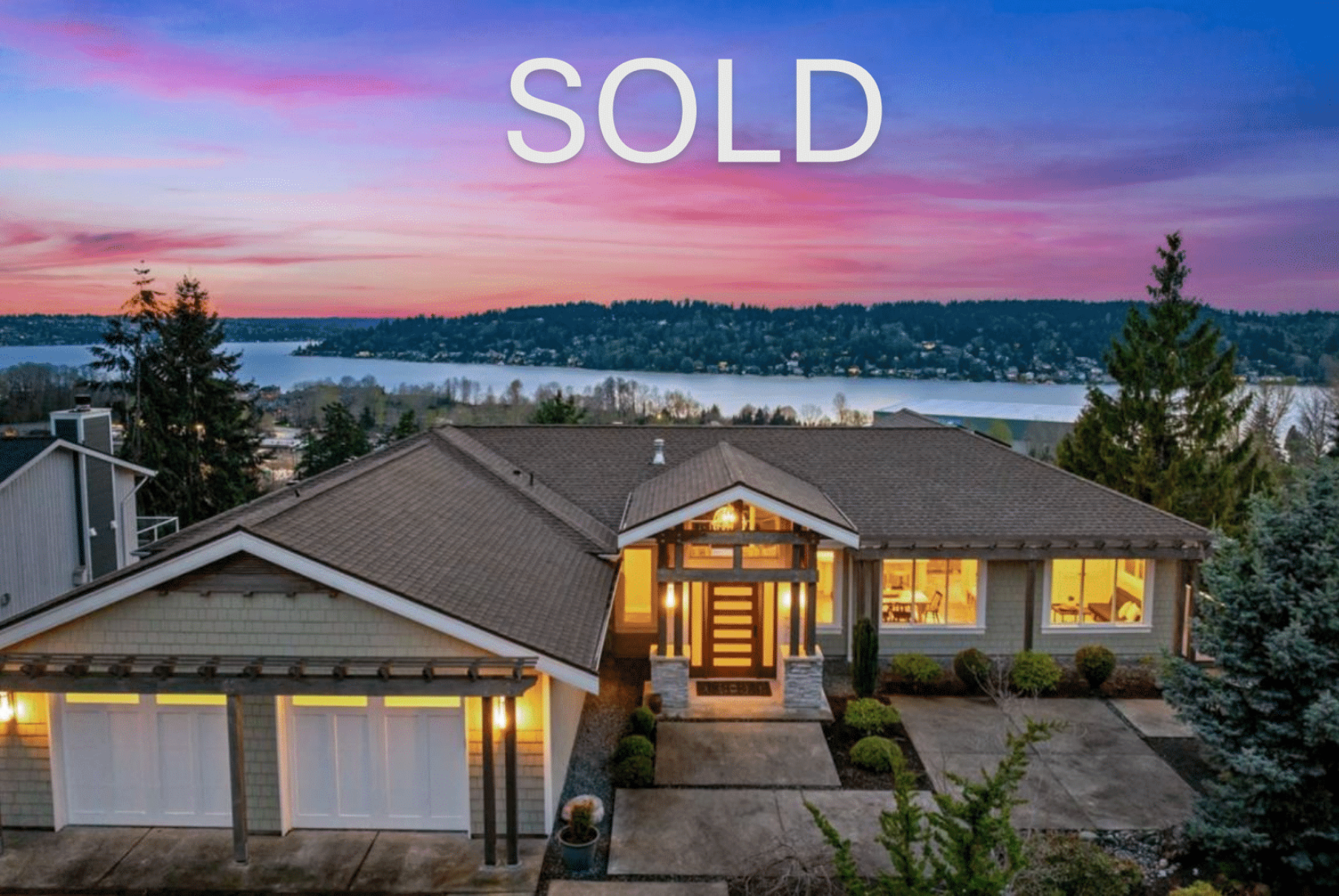 Sold sign on a modern house with large windows overlooking a scenic lake and sunset sky.