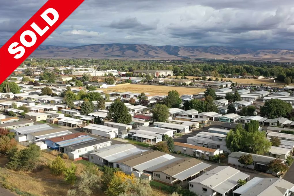 Aerial view of a suburban neighborhood with mobile homes and small houses, surrounded by green and dry fields, with mountains in the background, overlaid with the large blue and white text 