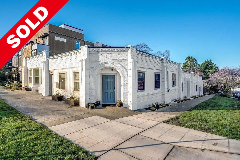 Sold sign on a white brick residential building in a suburban neighborhood with manicured lawn and flowering trees.