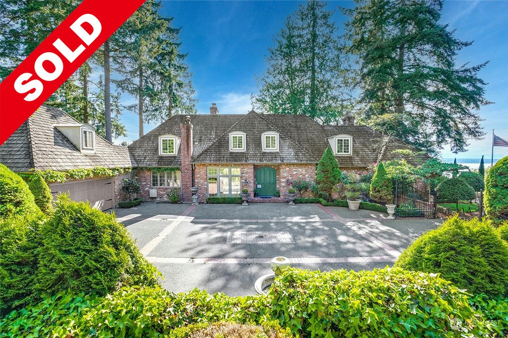 Sold sign on a house with a brick exterior, green door, and lush landscaping in a suburban setting with trees and a view of water in the background.
