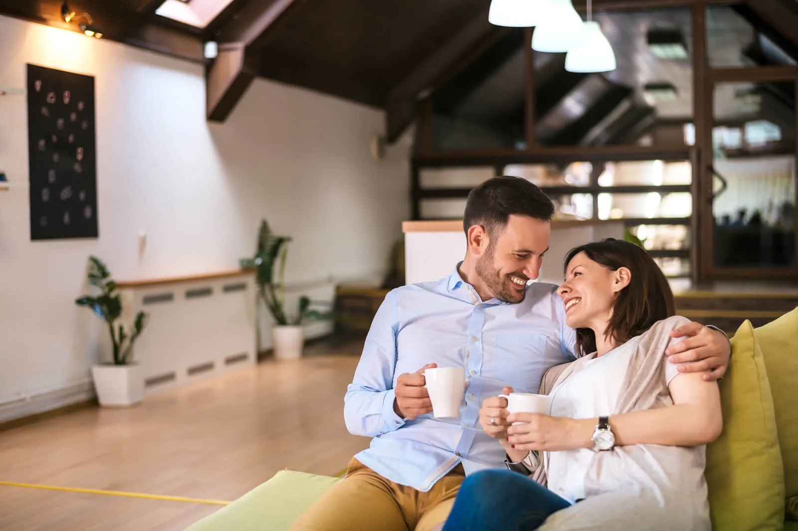 Smiling couple enjoying coffee together on a green sofa in a modern living room with wooden accents and indoor plants.