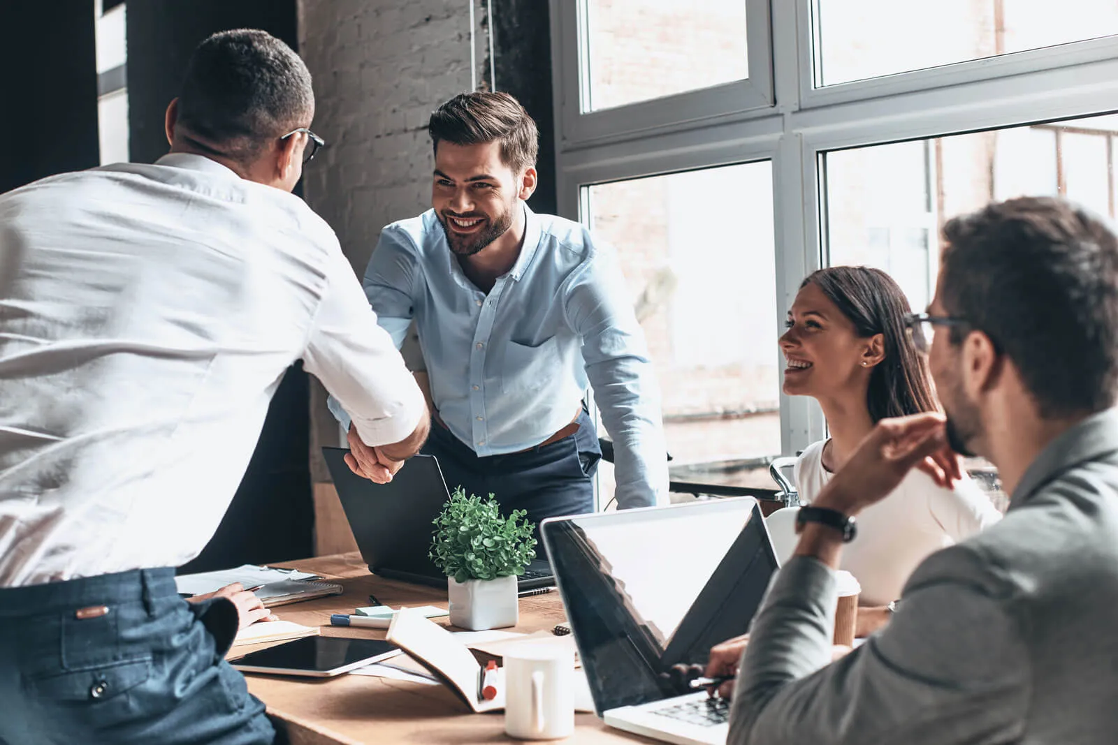Business team shaking hands in a modern office during daytime, smiling and collaborating with laptops and notebooks on the table.
