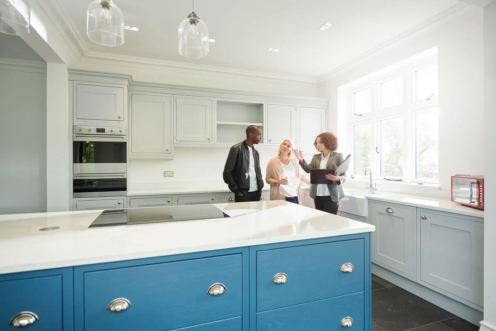 Real estate agents discussing a modern kitchen with large windows, white cabinets, and a blue island countertop.