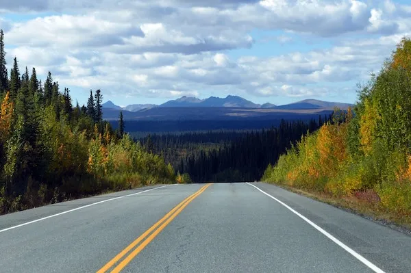 Scenic mountain road surrounded by autumn foliage and a forest, with mountains in the distance under a partly cloudy sky