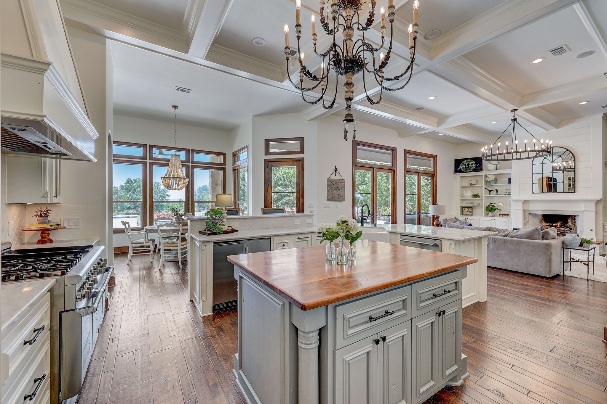 Spacious open-concept kitchen and living room with large windows, wooden flooring, elegant chandeliers, and cozy fireplace area.