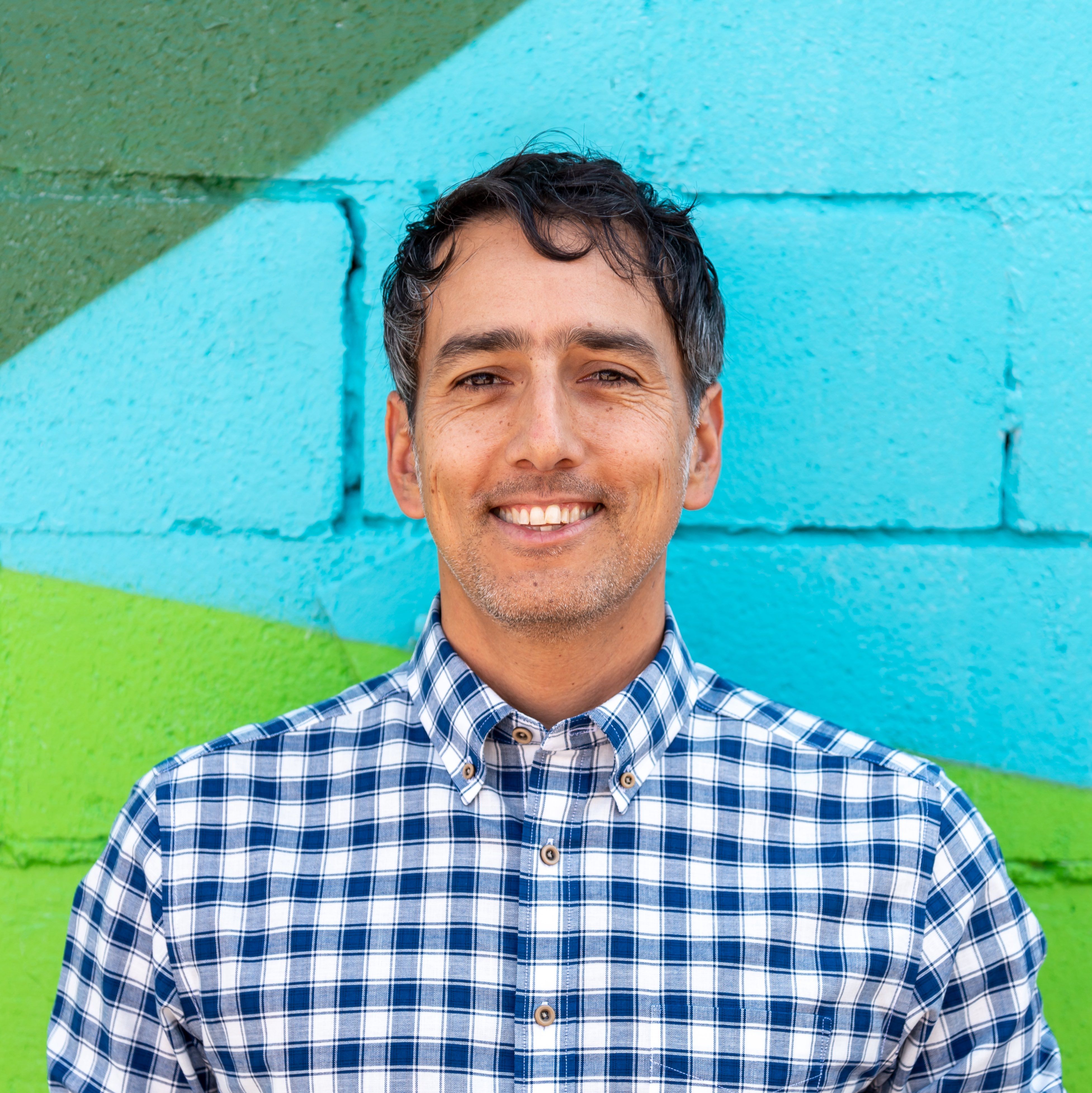Smiling man in checkered shirt standing in front of a colorful painted brick wall with blue and green sections.