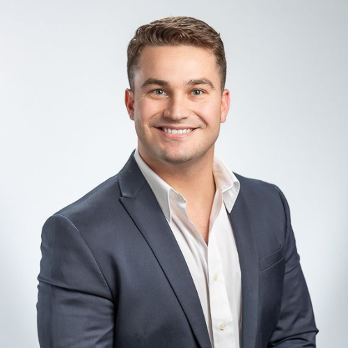 Professional portrait of a smiling young man in a navy blue suit and white shirt against a light gray background
