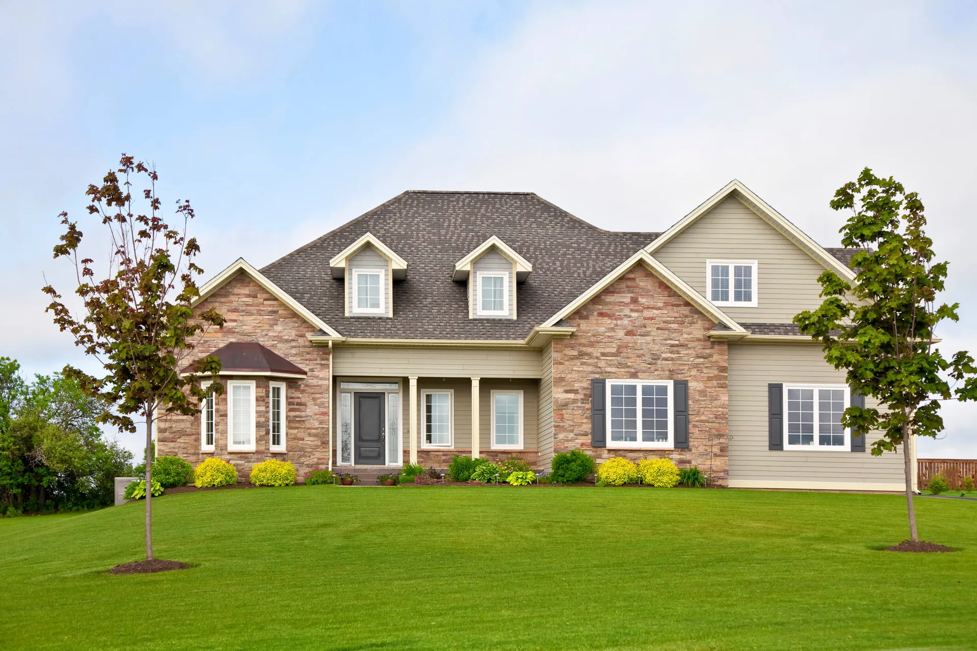 Spacious modern two-story house with beige siding, stone accents, black shutters, and a well-manicured lawn with two trees in front.