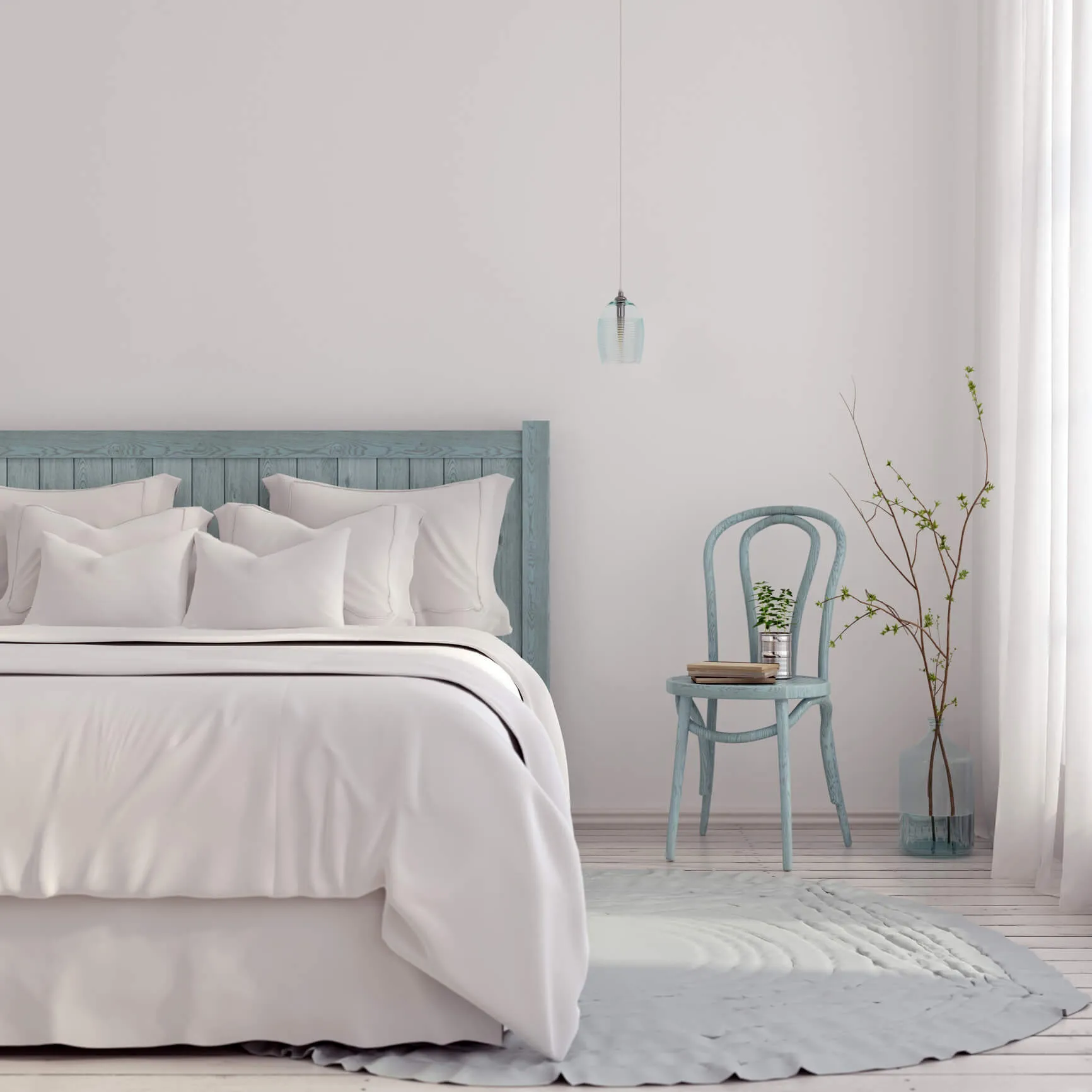 Cozy bedroom with a light blue wooden bed frame, white bedding, multiple pillows, a vintage blue chair with books and a potted plant, and a minimalist decor with a hanging pendant light, a tall vase with a branch, and white curtains letting in natural light.